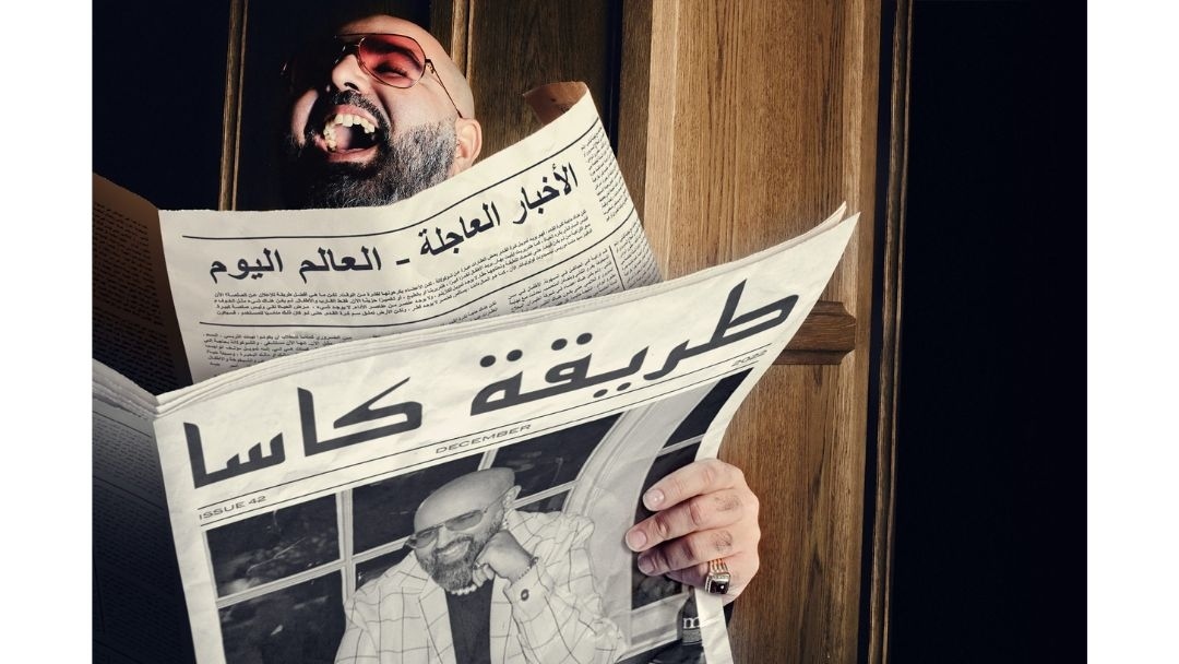 Man with glasses laughing behind two Arabic newspapers near a wood-paneled doorway, creating a humorous editorial portrait.
