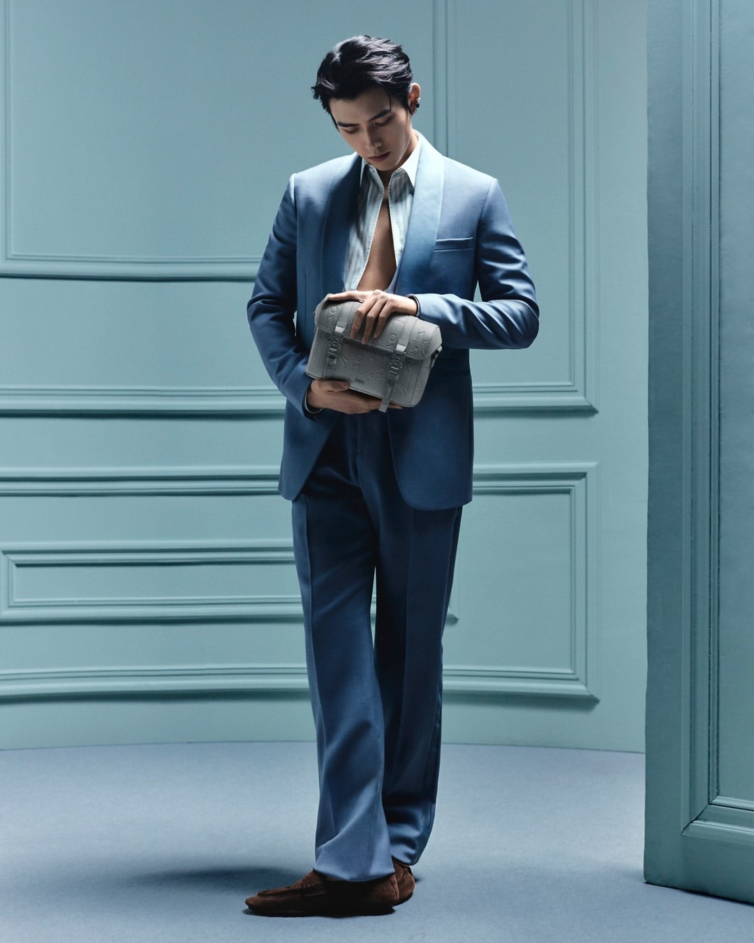 Man in a blue suit stands in front of a blue wall, holding a grey bag. He is looking down and wearing brown shoes.