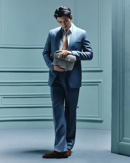 Man in a blue suit stands in front of a blue wall, holding a grey bag. He is looking down and wearing brown shoes.