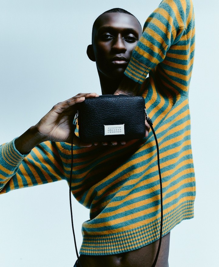 Adult male poses with black cross-body bag in a studio with soft lighting, wearing a striped sweater in a medium studio shot.