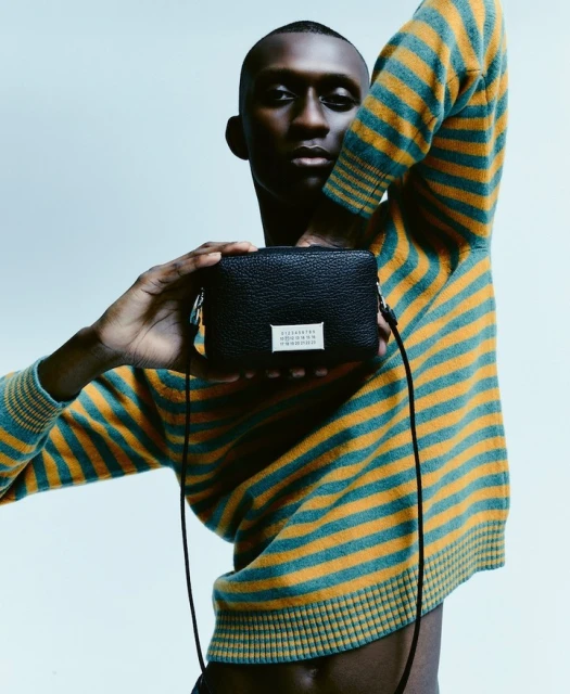 Adult male poses with black cross-body bag in a studio with soft lighting, wearing a striped sweater in a medium studio shot.