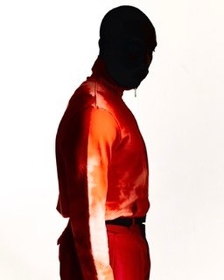 Profile shot of an adult wearing a red jumpsuit and a black ski mask, against a white backdrop, with a single light source.