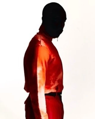Profile shot of an adult wearing a red jumpsuit and a black ski mask, against a white backdrop, with a single light source.