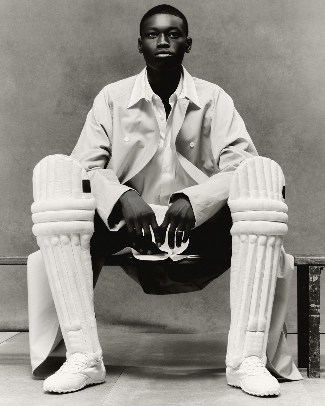 Portrait of a Black adult male seated wearing cricket pads, sneakers, shirt, and jacket in a photo studio with neutral background.