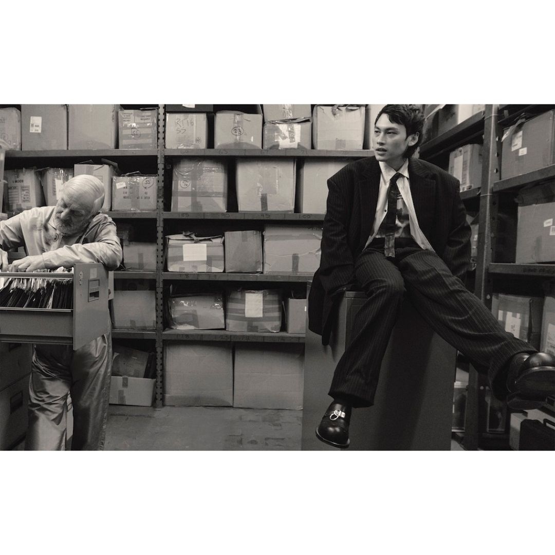 Monochrome shot of a stylish young man in suit sitting indoors, with an elderly man working amid shelves of cardboard boxes.