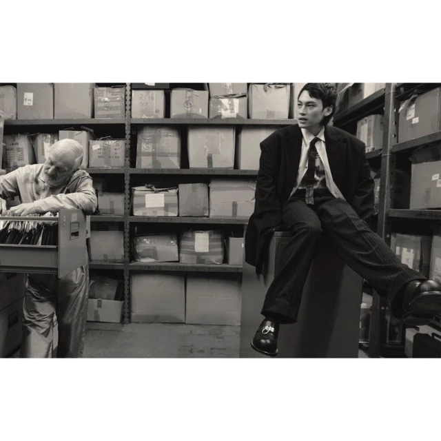 Monochrome shot of a stylish young man in suit sitting indoors, with an elderly man working amid shelves of cardboard boxes.