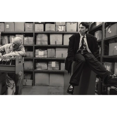 Monochrome shot of a stylish young man in suit sitting indoors, with an elderly man working amid shelves of cardboard boxes.