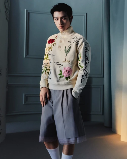 Adult male fashion model poses in an eye-level medium shot, wearing a flower-embroidered sweater, grey shorts, and white socks in an indoor studio.