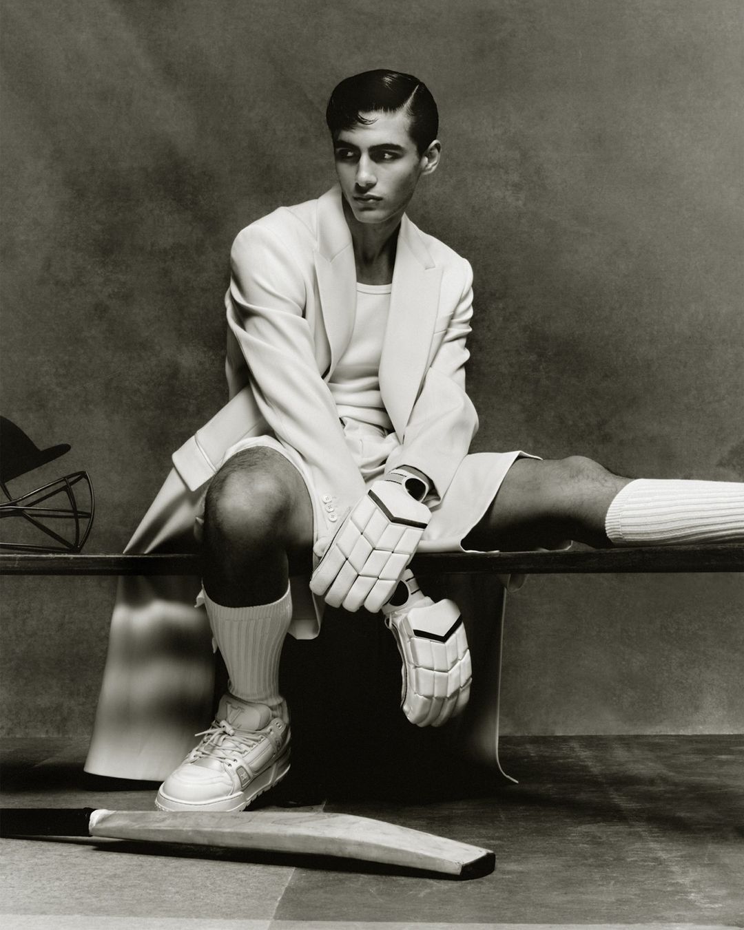 Monochrome portrait of a man in sports attire sitting on a bench with cricket equipment, creating a vintage athletic fashion statement.