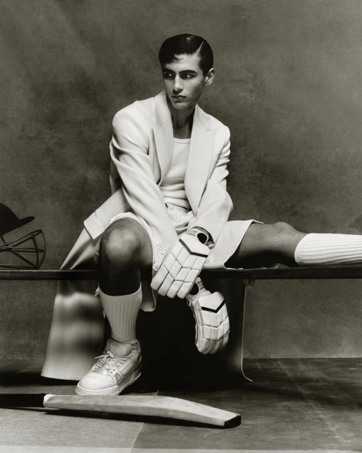 Monochrome portrait of a man in sports attire sitting on a bench with cricket equipment, creating a vintage athletic fashion statement.