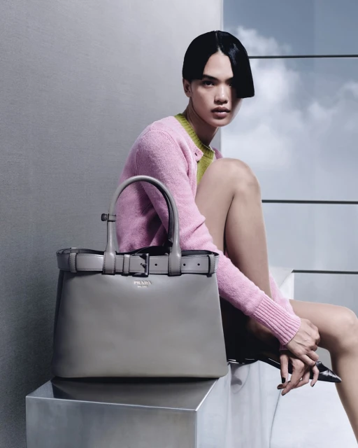 A woman in a pink cardigan poses with a grey leather Prada handbag, seated in an indoor fashion studio setting with natural lighting.
