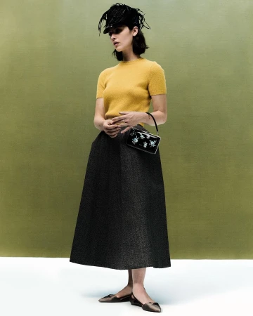 Model in a yellow sweater and dark midi skirt wearing a feathered cap and holding a small handbag