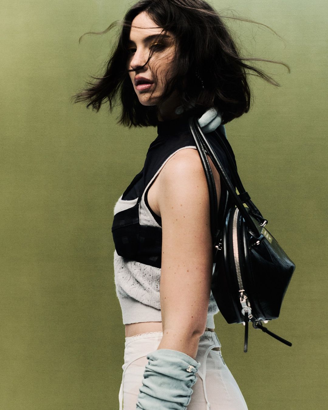 Portrait of a woman in a sleeveless top, sheer skirt, and blue gloves with a black bag, standing against a green background.