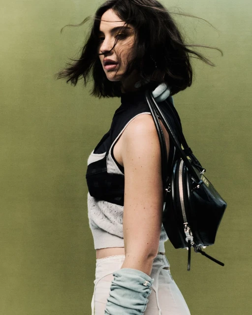 Portrait of a woman in a sleeveless top, sheer skirt, and blue gloves with a black bag, standing against a green background.