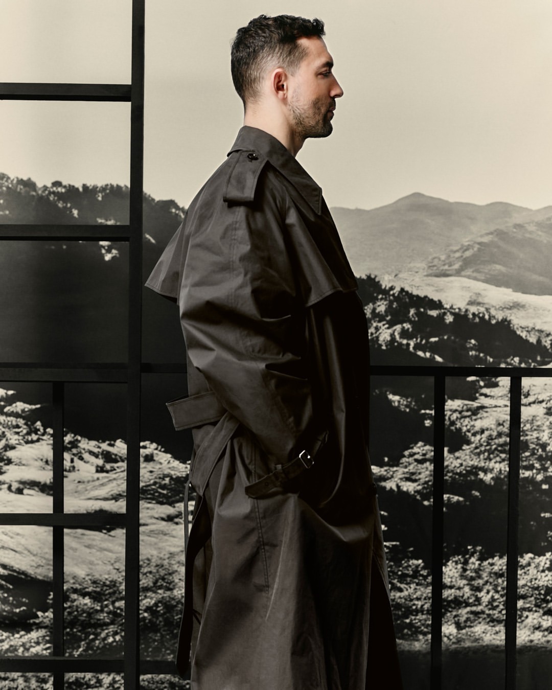 Man in a black trench coat stands in profile against a backdrop of mountains, framed by a window grid. Fashion editorial.