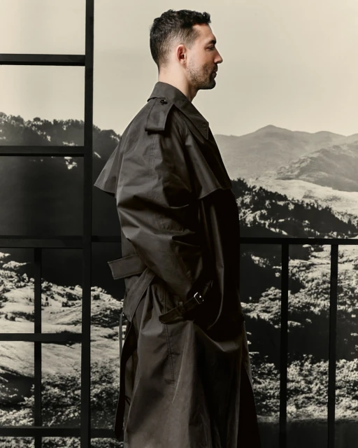 Man in a black trench coat stands in profile against a backdrop of mountains, framed by a window grid. Fashion editorial.