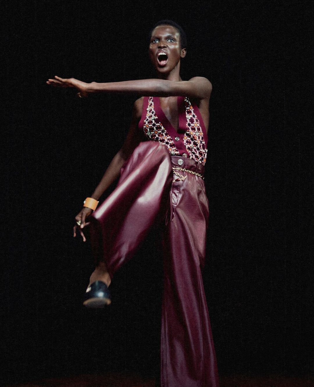 Woman in performance art pose wearing maroon pants and jeweled top, arm outstretched against dark backdrop in a studio.