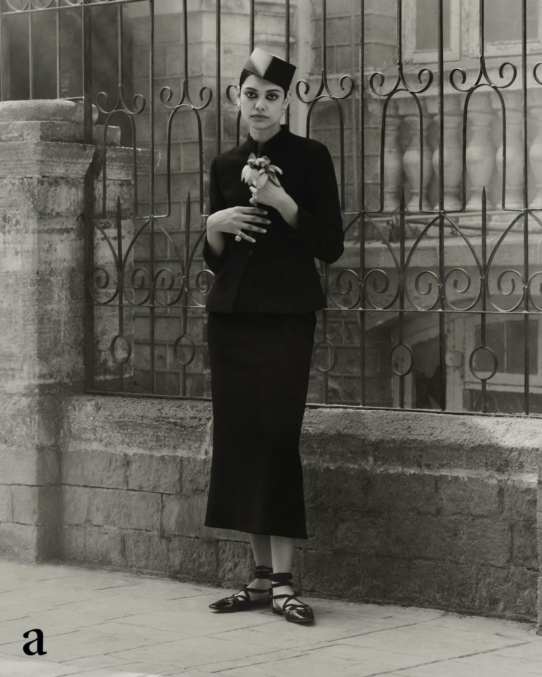 Woman in black dress stands near an iron gate holding a flower, photographed in black and white with shallow depth of field.