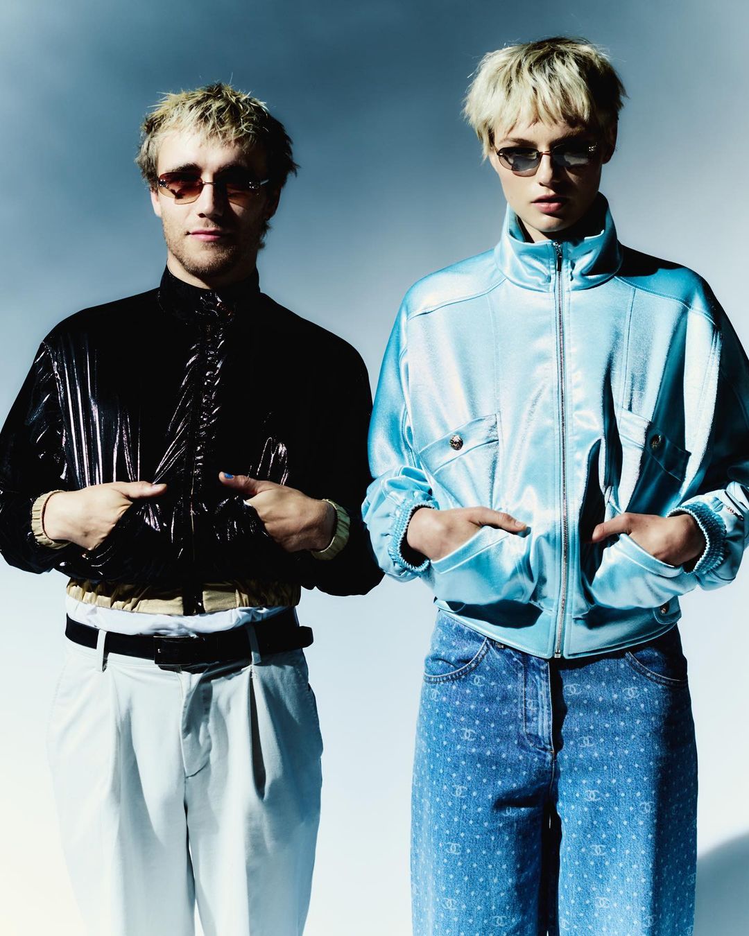 Two fair-skinned people with blonde hair pose indoors. They wear sunglasses and shiny jackets, and jeans. A minimalist studio background.