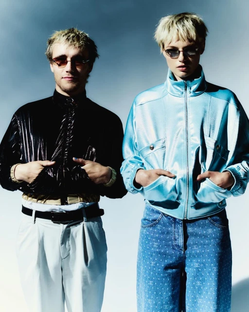 Two fair-skinned people with blonde hair pose indoors. They wear sunglasses and shiny jackets, and jeans. A minimalist studio background.