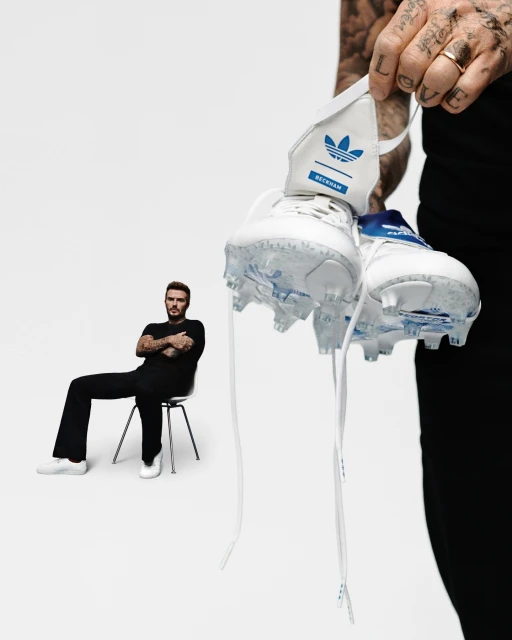 David Beckham sits in the background as white and blue Adidas soccer shoes are held up on a white backdrop in a studio shot.