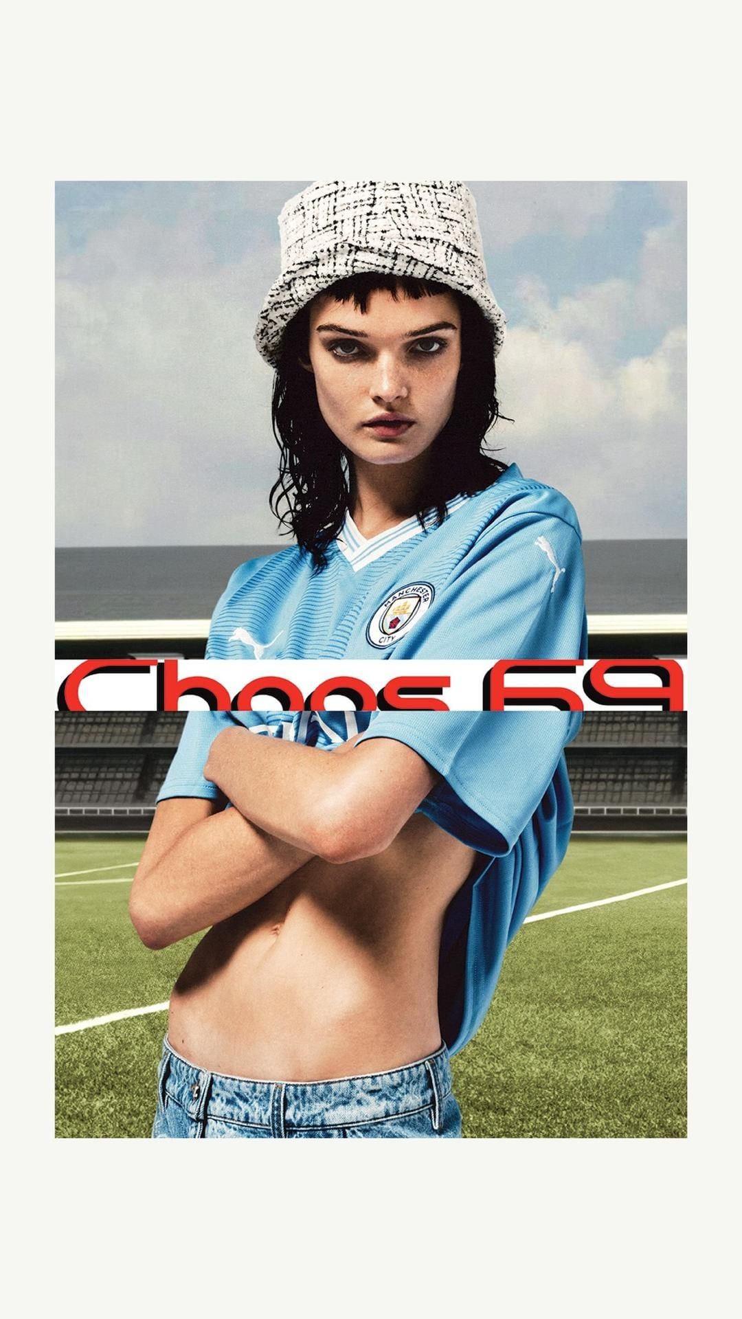 Fashion shot of a woman in a blue jersey and denim shorts, wearing a bucket hat, posing outdoors on a sports field, with stadium seating.