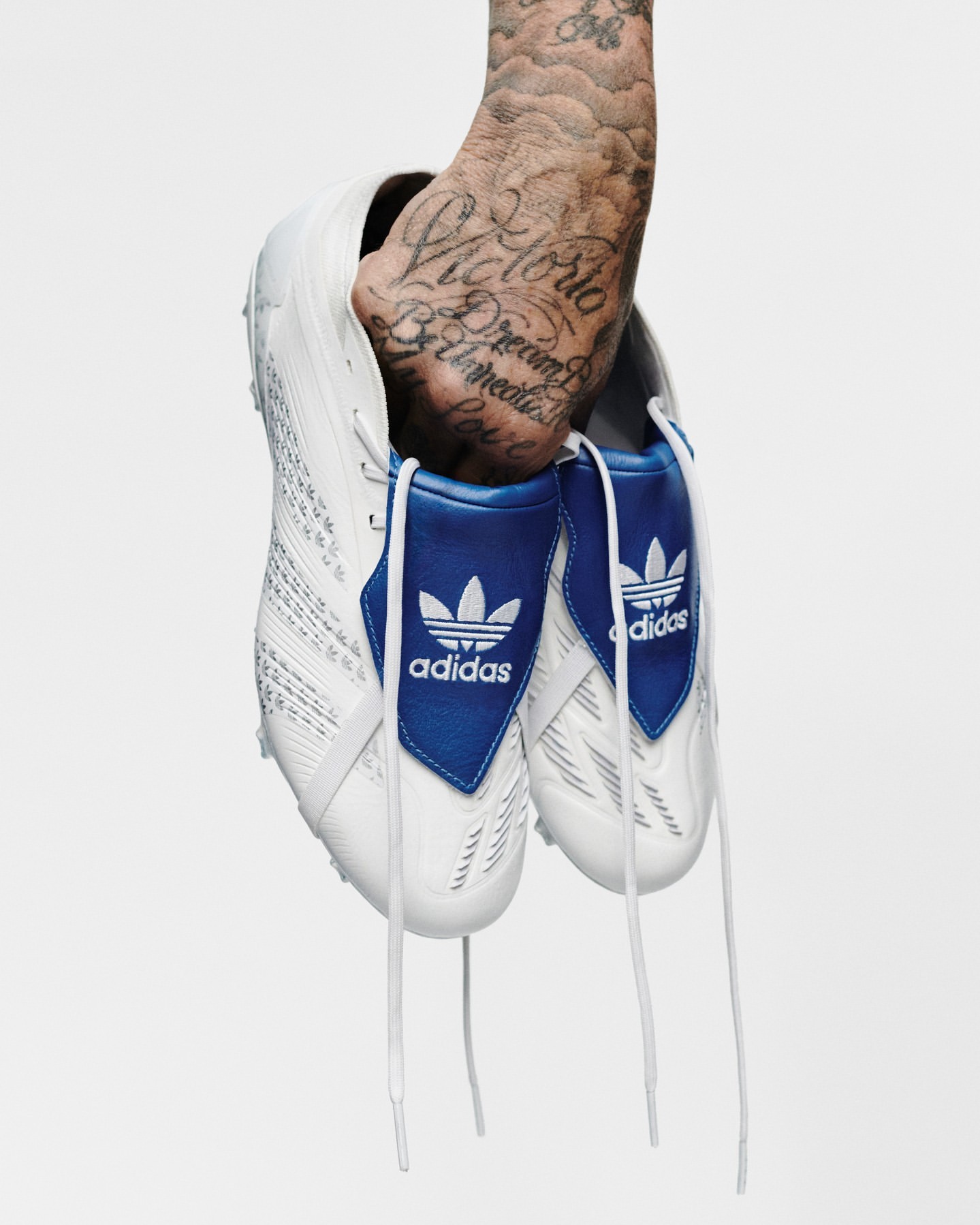 Hand holding pair of white Adidas soccer cleats with blue trim and laces against a white background; arm has visible tattoos.