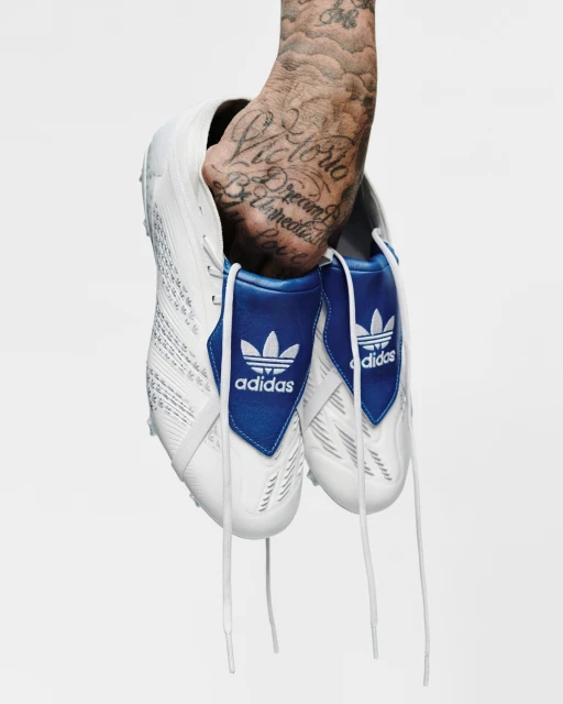 Hand holding pair of white Adidas soccer cleats with blue trim and laces against a white background; arm has visible tattoos.