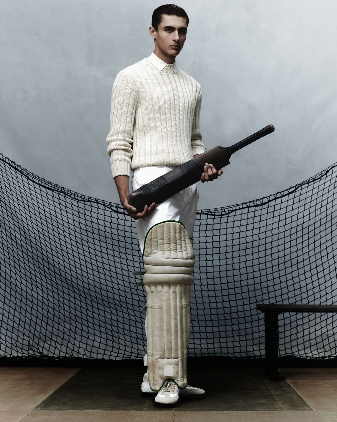 An adult male is posing indoors wearing sports clothing while holding a cricket bat. Leg pads and a black net are visible.