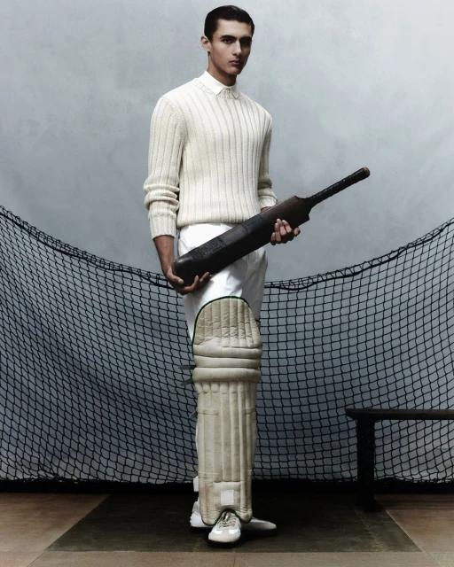 An adult male is posing indoors wearing sports clothing while holding a cricket bat. Leg pads and a black net are visible.