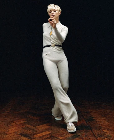 Blonde woman wearing a cream knitted jumpsuit and white sneakers posing against a dark herringbone floor.