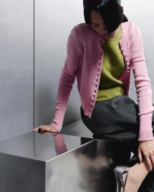 Woman in pink cardigan sitting on a reflective metallic surface, wearing a green top and dark skirt, with a neutral grey background.