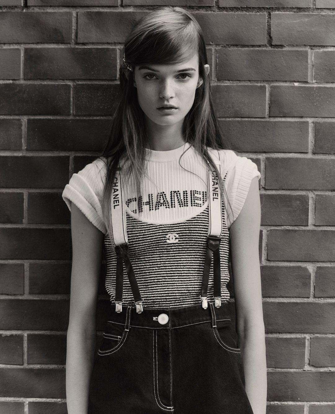 Monochrome portrait of a young adult woman in a Chanel outfit against a brick wall, eyes forward, in a fashion editorial style.