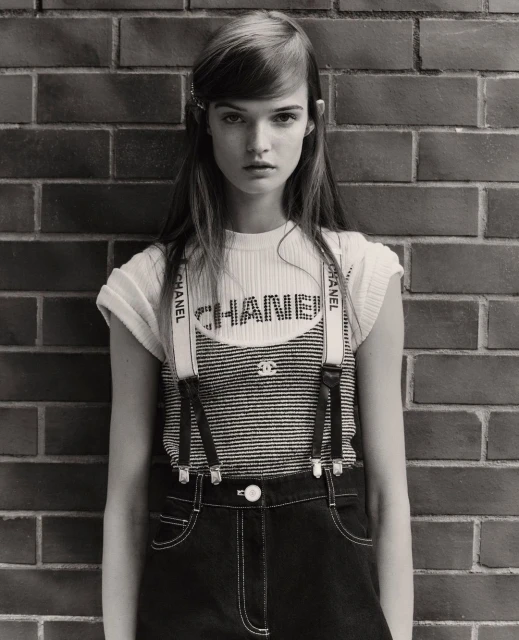 Monochrome portrait of a young adult woman in a Chanel outfit against a brick wall, eyes forward, in a fashion editorial style.