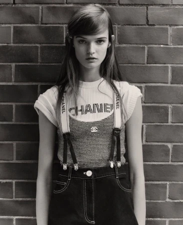 Monochrome portrait of a young adult woman in a Chanel outfit against a brick wall, eyes forward, in a fashion editorial style.