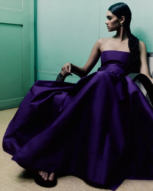 An adult female sits in a chair wearing a strapless violet gown with a braided hairstyle against an indoor aqua-colored wall.