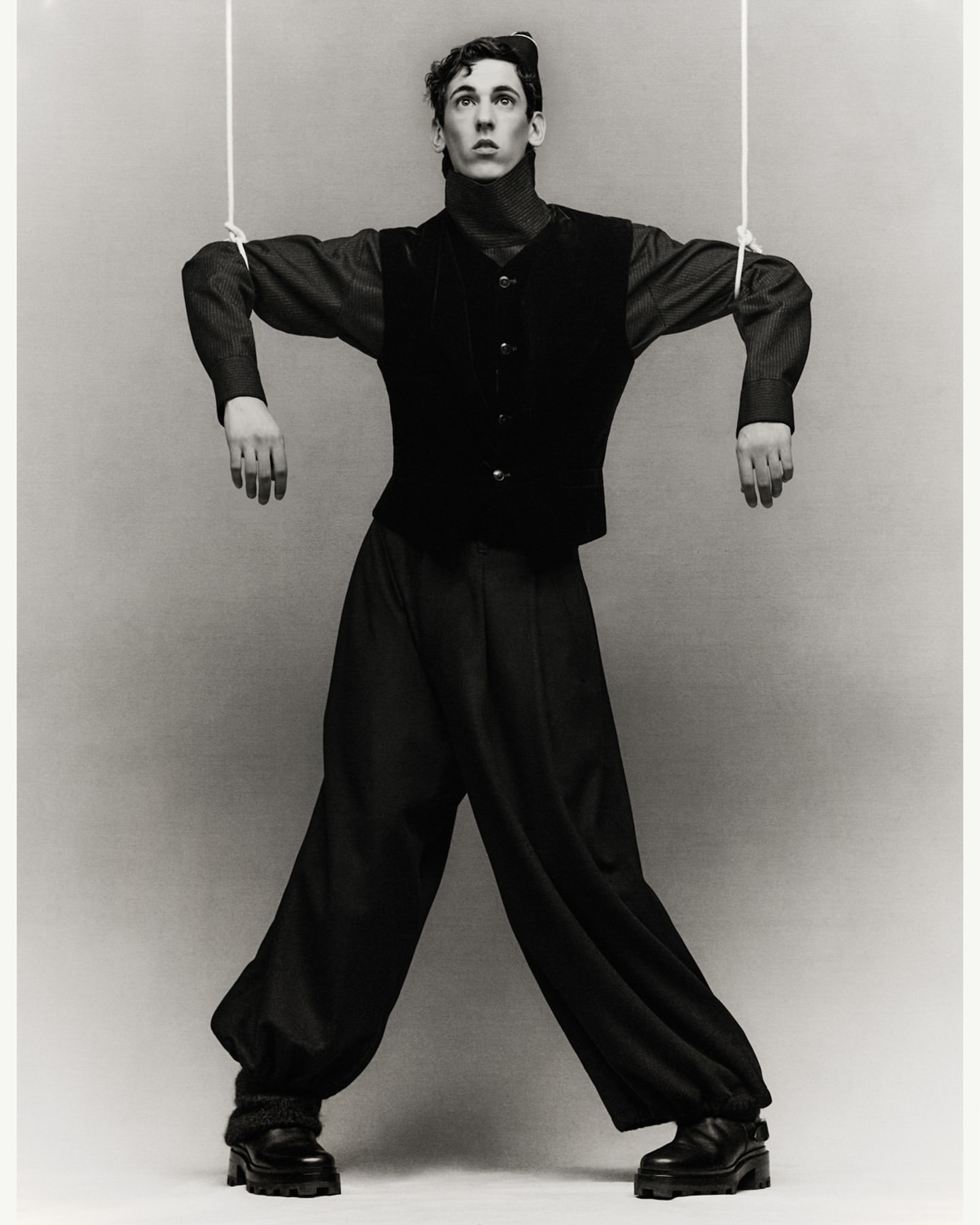 Monochrome image of a fair-skinned male in a dark vest and pants, mimicking a puppet, with strings attached to his arms and head.