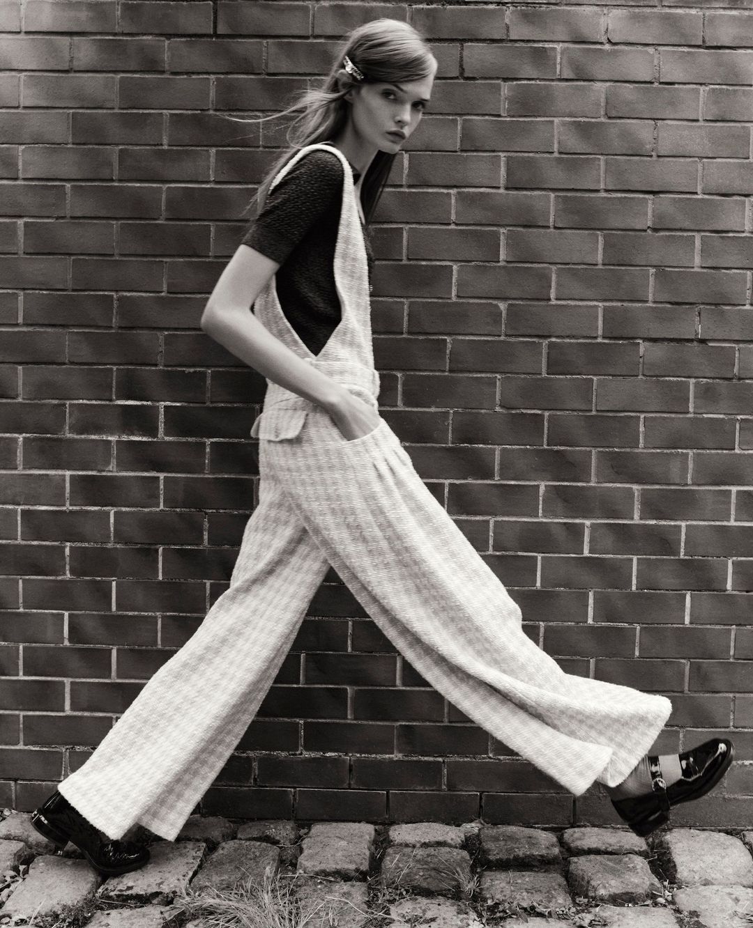 Black-and-white fashion shot of a slender model in overalls walking against a brick wall, showcasing retro-inspired style and design.