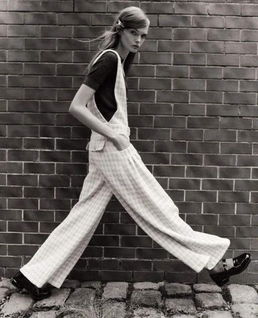 Black-and-white fashion shot of a slender model in overalls walking against a brick wall, showcasing retro-inspired style and design.