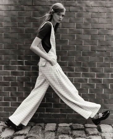 Black-and-white fashion shot of a slender model in overalls walking against a brick wall, showcasing retro-inspired style and design.