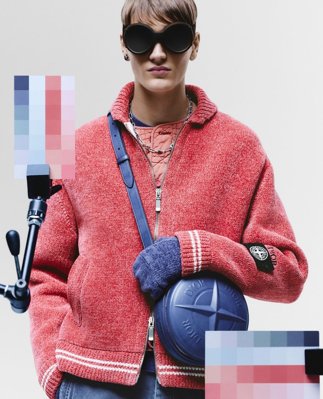 Fashionable individual with sunglasses poses indoors, wearing a red jacket, carrying a blue round purse, and standing against a neutral backdrop.