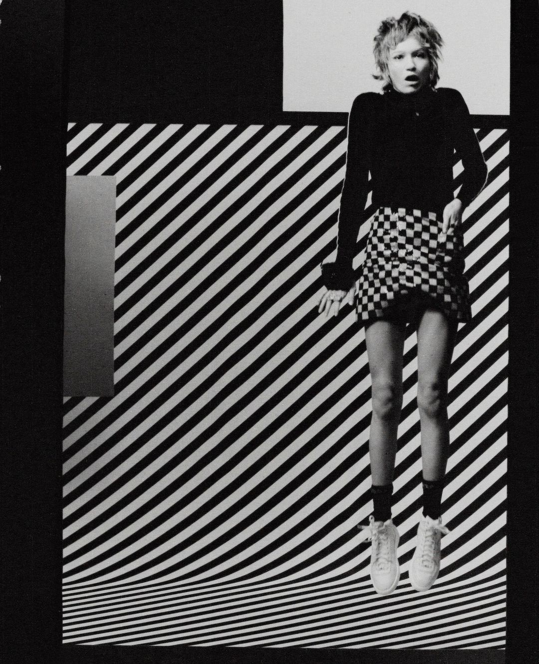 Monochrome fashion shot of a fair-skinned female adult mid-air wearing checker-print skirt, top, sneakers against striped backdrop.