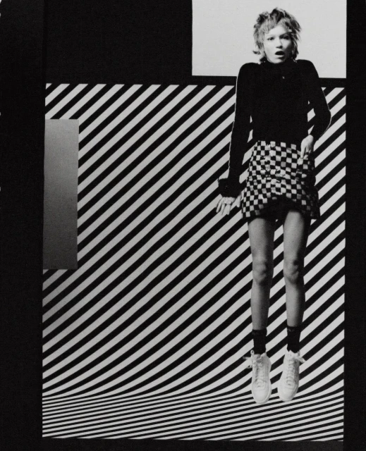 Monochrome fashion shot of a fair-skinned female adult mid-air wearing checker-print skirt, top, sneakers against striped backdrop.