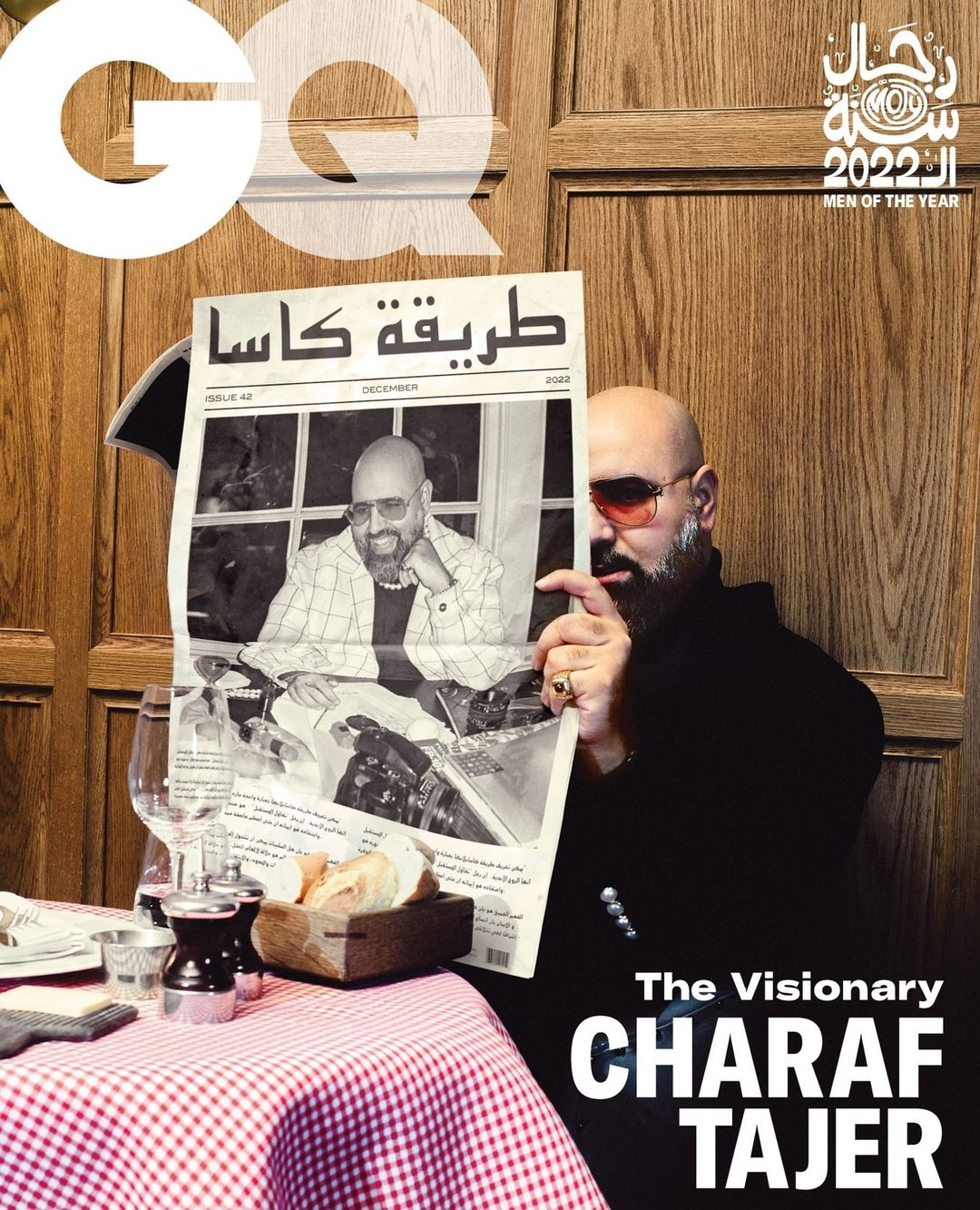 Man holding newspaper with his own photo on GQ magazine cover in restaurant setting, featuring checked tablecloth and glassware.