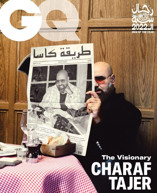 Man holding newspaper with his own photo on GQ magazine cover in restaurant setting, featuring checked tablecloth and glassware.