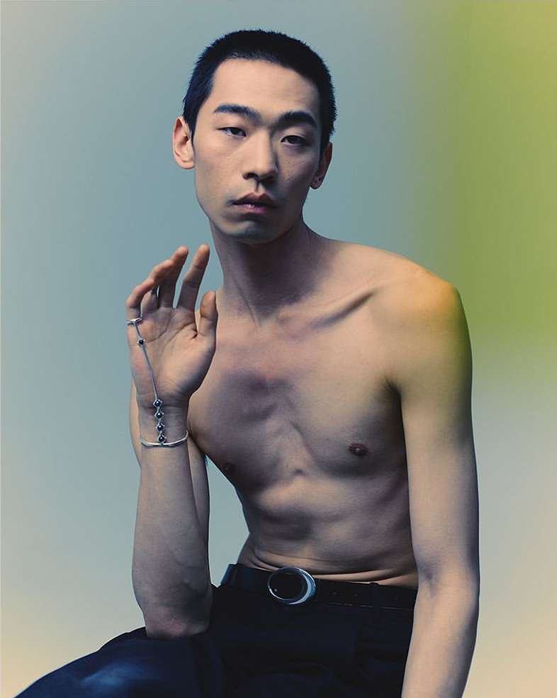 A shirtless Asian male model with short hair poses seated indoors, wearing black pants and a silver hand bracelet in a portrait.