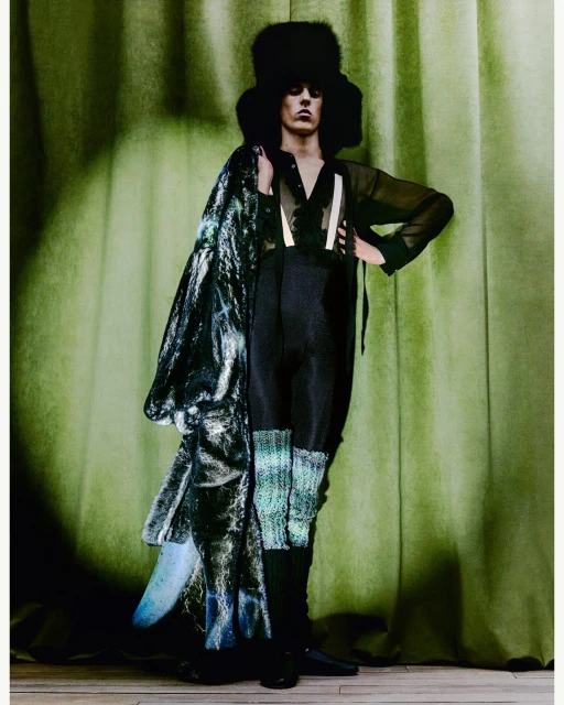 A person in dark clothing and a large hat poses in front of a green curtain, holding a coat with a shiny blue-green pattern.
