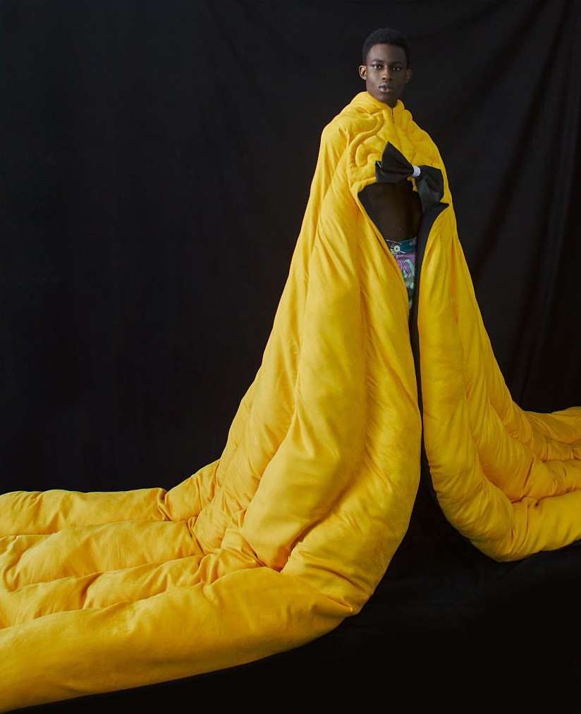 Fashion shot of a Black male model draped in a voluminous yellow quilted garment with a black bow, posing against a black backdrop.