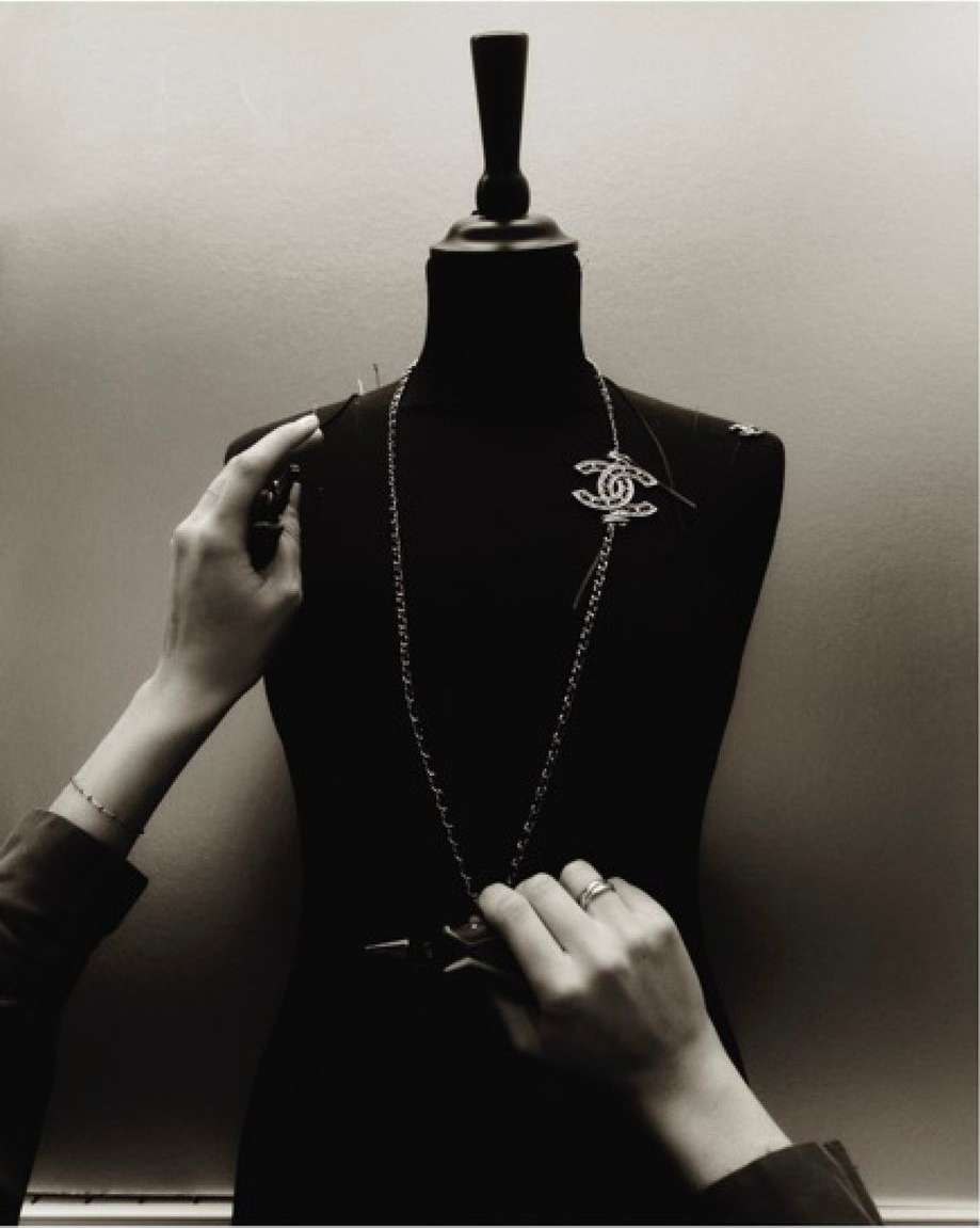Black and white studio shot of a mannequin being prepared for a fashion shoot with hands pinning a Chanel brooch.
