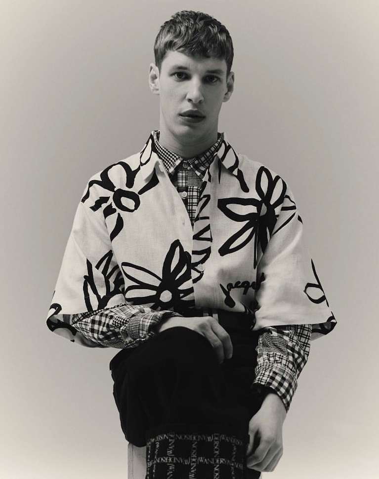 Monochromatic portrait of a young adult male model wearing a floral print shirt, plaid shirt, and dark pants in a light studio setting.